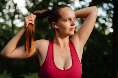 Kırmızı atletik bluzlu kararlı bir kadın güneşli bir parkta ya da ormandaki spor yaparken uzun saçlarını at kuyruğuna takıyor ve kulaklık takıyor..