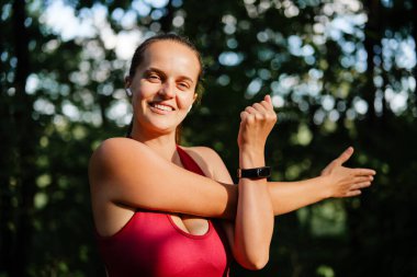 Kırmızı atletik bluzlu, spor ayakkabılı, kulaklıklı, güler yüzlü bir kadın güneşli bir ormanda kol ve omuz hareketleri yapıyor..