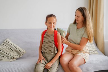 Kırmızı sırt çantalı, gülümseyen okul çağındaki bir kız, annesi yanına oturmuş, okul hazırlıkları için ders kitaplarını ya da defterleri çantaya koyarken koltuğa oturur..