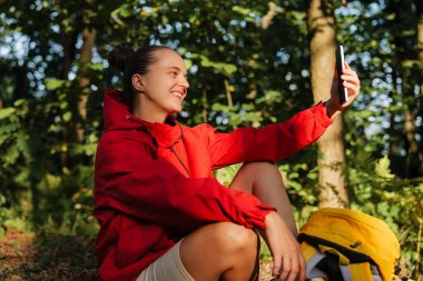 Genç bir kadın, yeşil ve güneş ışığıyla çevrili telefonuyla selfie çekerken gülümseyerek bir ormanda yerde oturuyor..