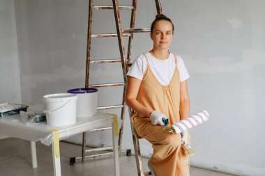 Kısmen boyanmış bir odada merdivende oturan bir kadın elinde boya silindiri ile başlamaya hazır görünüyor. Boya malzemeleri yakınlarda, tadilat yapıldığını gösteriyor..