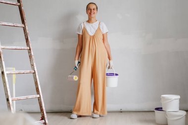 Yeni boyanmış bir odada elinde boya aletleri olan ve kovalarla çevrili neşeli genç bir kadın ev iyileştirme görevi için hazır bekliyor..