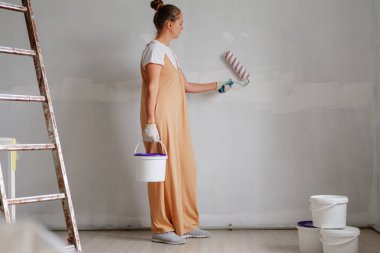 Bir kadın uzun bir önlük giyerken duvara silindirle boya sürüyor. Bir merdivenin yanında duruyor ve diğer elinde bir kova boya tutuyor..