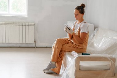 Tulum giymiş bir kadın kanepeye oturur ve etrafı kısmen yenilenmiş bir odayla çevriliyken telefonuna odaklanır..