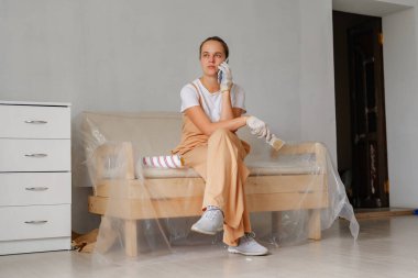 Gündelik giysiler içinde genç bir kadın ahşap bir koltukta oturuyor, elinde fırçayla bir oda yenileme sırasında telefonla konuşuyor..