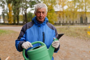Mavi ceketli yaşlı bir adam yeşil bir sulama tenekesi tutuyor, sıcak sonbahar güneşi ve renkli yaprakların tadını çıkarırken bir parkta bitkilerle ilgileniyor..