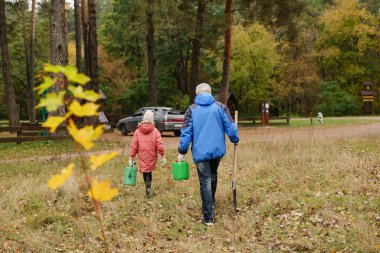 Huzurlu bir ormanda, bir adam ve bir çocuk yeşil kovaları ve renkli sonbahar yaprakları arasında bahçe aletlerini tutarak çimenli bir yolda yürürler..