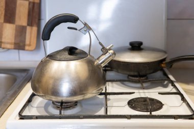 Parlak metal bir çaydanlık gaz sobasının üzerinde duruyor. Kızartma tavası yakınlarda, gündüz vakti aydınlık bir mutfakta rahat bir sahne oluşturuyor..