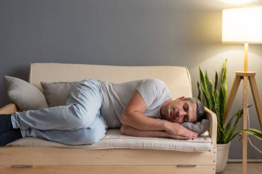 Yumuşak bir kanepede uyuyan bir adam sıcak bir oturma odasında, bitkilerle ve yumuşak ışıklarla çevrili, dinlenmek için rahatlatıcı bir atmosfer yaratıyor..