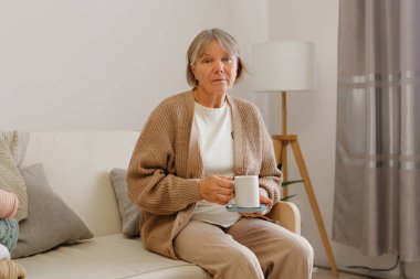 Gri saçlı olgun bir kadın elinde bir fincan çayla açık bir kanepeye oturur ve parlak, davetkar bir oturma odasında düşünceli bir şekilde bakar..
