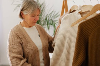 Bir kadın, yeşilliklerle çevrili bir butikte, yumuşak süveterleri tarafsız bir tonda inceler ve öğleden sonraları rahat bir alışveriş deneyimi yaşar..