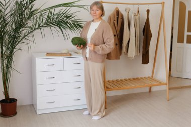 Bir kadın, sıcak bir odada asılı giysiler ve şifoniyerin yanında uzun bir bitki ile dururken yeşil iplik tutar..