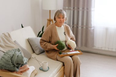 Son sınıf öğrencisi bir kadın, elinde yeşil ve beyaz ipliklerle, parlak bir oturma odasında, kanepede rahatça oturup örgü örmeye hazırlanıyor. Odaklanmış ve memnun görünüyor..