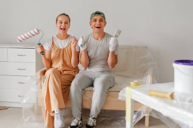 Neşeli bir çift, oturma odalarını boyamaya hazır plastikle kaplı bir kanepede oturur. Ellerinde boya malzemeleri var ve ev projeleri için çok heyecanlılar..