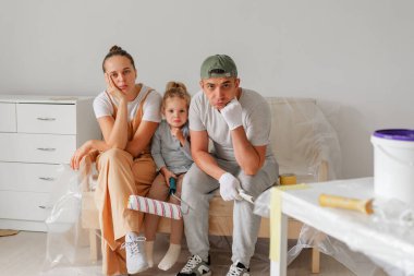 Bir aile evlerinin tadilatına ara veriyor. Çift ve küçük kızları boya malzemeleriyle çevrili bir kanepede otururken yorgun görünüyorlar..