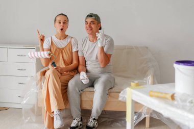 Mutlu bir çift, oturma odasının şeklini değiştirmekten heyecan duyan boya aletleriyle bir koltukta oturur. Çevrelerinde eğlenceli bir atmosfer ve boya malzemeleri var..