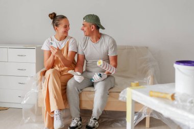 Genç bir çift, güneşli bir haftasonunda, parlak ve yeni hazırlanmış bir odayla çevrili bir kanepede oturup gülümseyip boya aletleri tutarlar..