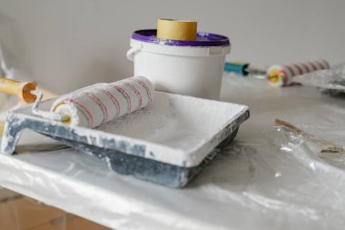 Kovalar ve silindirler plastik kaplamayla kaplı bir masanın üzerine yerleştiriliyor, iyi ışıklandırılmış bir mekandaki ev yenileme sırasında boyamak için çalışma alanını gösteriyor.