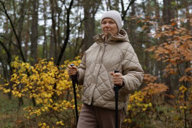 Bej ceketli ve beyaz şapkalı yaşlı bir kadın huzurlu bir ormanda duruyor, elinde güzel sonbahar yaprakları arasında yürüyüş direkleri tutuyor..