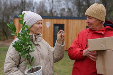 İki kişi, serin bir sonbahar öğleden sonrasında ahşap bir barakanın yanında halk bahçesinde dururken bahçe aletlerini ve küçük bir bitkiyi değiş tokuş ediyorlar..