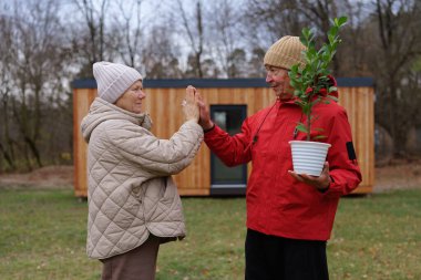 Kırmızı ceketli bir adam ve yorgan giymiş bir kadın, sonbaharda bahçelerine küçük bir ağaç dikmeye hazırlanırken neşeli bir anı paylaşıyorlar..