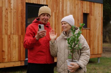 Yaşlı bir çift, ahşap evlerinin dışındaki bir video çağrısı sırasında gülümseyip etkileşime geçiyor, serin bir sonbahar gününde saksı bitkisi tutuyor..