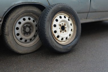 Islak bir yolda birbirine yakın duran iki araba tekerleği, birinde belirgin bir aşınma, diğerinde ise hasar ve yaş belirtileri var..