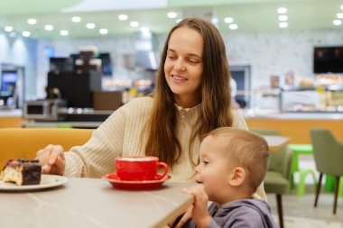 Bir anne, küçük çocuğu karşılama kafesinde tatlıya bakarken kahvesini yudumlar. Sakin bir öğleden sonra bir anlık neşeyi paylaşıyorlar..