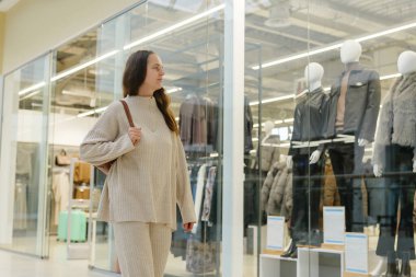 Açık renk süveter ve pantolon giyen bir kadın mağaza vitrinlerinde sergilenen kışlık kıyafetlere hayran bir şekilde çağdaş bir alışveriş merkezinde geziniyor..