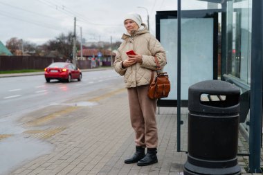 Bir kadın otobüs durağında duruyor, örtülü ceket ve şapka giymiş. O bir telefon tutar ve sabırla caddede arabalar geçerken bekler..