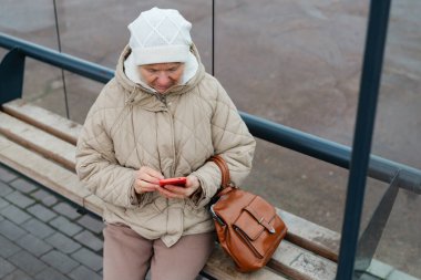 Sıcak bir palto ve şapka giyen yaşlı bir kadın otobüs durağındaki bankta oturuyor, telefonuna odaklanmış, serin bir günde binmesini bekliyor..