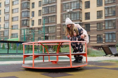 Bir anne, çocuğunun modern apartmanlarla çevrili bir oyun parkında atlıkarıncaya binmesine yardım eder. Güneşli öğleden sonraları keyifli anları paylaşıyorlar..