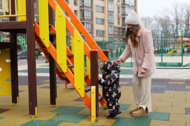 Bir anne, serin havalarda şehir parkında sakin bir günün tadını çıkarırken çocuğunun renkli bir oyun parkında merdivenleri tırmanmasına yardım eder..