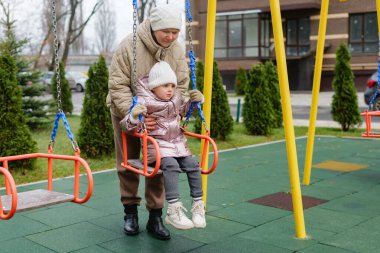 Bir büyükanne, serin bir günde torununu salıncakta sallıyor. Ağaçlar ve şehir arkaplanı yakınlarda görülebilir.