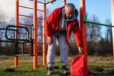 Kıdemli bir adam, açık havada oyun alanı ekipmanlarının yanında egzersiz yapmayı sever. Açık havada kırmızı sırt çantasına uzanmak için bir dakikasını ayırır..