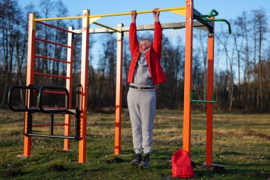 Son sınıf öğrencisi, parlak öğleden sonra ışığında ağaçlarla çevrili sakin bir parkta renkli spor aletleriyle barfiks çekiyor..