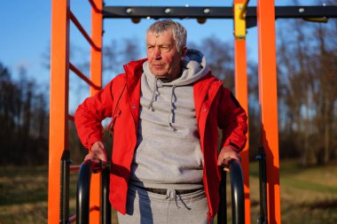 Kırmızı ceket ve gri eşofman giyen son sınıf öğrencisi açık hava spor salonunda güneşli bir kış öğleden sonrasında parkta dikiliyor..