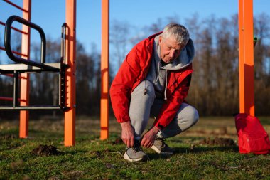 Kırmızı ceket giyen yaşlı bir adam ayakkabı bağcıklarını dışarıdaki spor aletlerinin yanına bağlamak için eğiliyor, yeşil bir parkta açık bir sabahın tadını çıkarıyor..