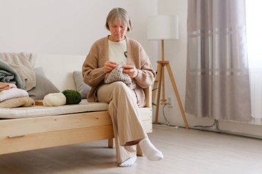 Bir kadın, iyi aydınlatılmış bir oturma odasında rahat bir kanepede örgü örmekten hoşlanır. Yumuşak iplik ve rahat bir atmosfer el işi için sakin bir ortam yaratır..