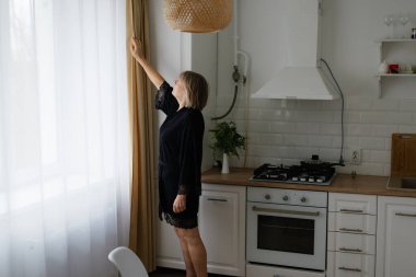 Siyah bornozlu bir kadın güneşli bir mutfakta perde çekiyor ve doğal ışığın boşluğu doldurmasına izin veriyor. Bitkiler ve soba olay yerine sıcaklık katar..