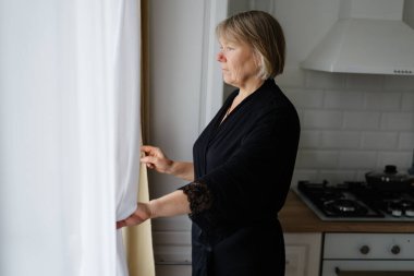 Modern bir mutfağın penceresinde siyah bir bornoz giymiş bir kadın duruyor. Dışarıya bakıyor, gündüz saatlerinde içeri giren doğal ışığın tadını çıkarıyor..