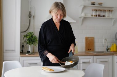 Siyah bornozlu bir kadın, parlak bir mutfakta duruyor. Sabah yemeğini hazırlarken kızarmış yumurtayı tavadan tabağa ustalıkla çeviriyor..