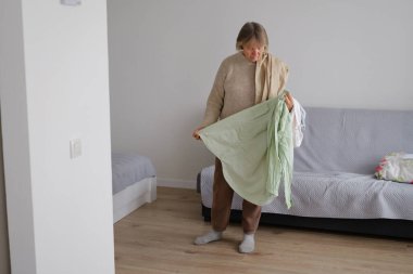 Bir kadın minimalist bir odada duruyor, güne hazırlanırken açık yeşil bir gömleği inceliyor. Yumuşak ışık boşluğu aydınlatır.