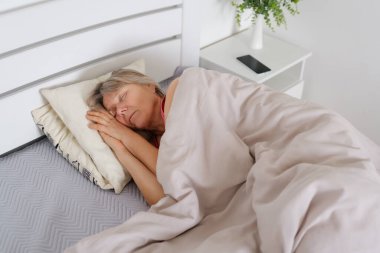 Yaşlı bir kadın, sakin ve düzenli bir yatak odası atmosferiyle çevrili yumuşak battaniyelere sarılı yatağında huzurlu bir uyku çeker..