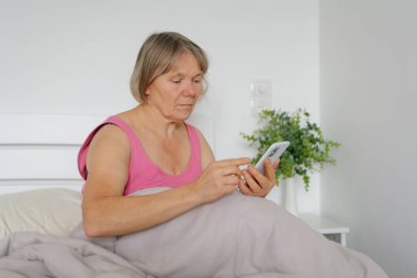 Kadın yatakta oturmuş, akıllı telefonuna odaklanmış. Yumuşak bir battaniyeye sarılmış, muhtemelen evde sakin ve rahatlatıcı bir sabahın tadını çıkarıyor..