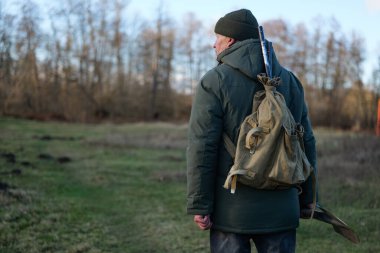 Sırt çantası ve bastonuyla yaşlı bir adam, ilkbaharın başlarında açık mavi bir gökyüzünün altında ağaçlar ve çimlerle çevrili bir orman yolunda yürüyor..