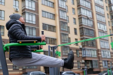 Kışlık ceketli bir adam, serin bir günde, uzun apartman binalarıyla çevriliyken spor salonunda spor yapıyor..