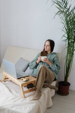 Bir kadın, dizüstü bilgisayarını kullanırken sıcak bir içkinin tadını çıkarırken koltukta rahatça oturur. Etrafı doğal ve sakin bir atmosferle çevrili..