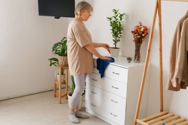 Bej gömlekli bir kadın, mavi bir kumaşı beyaz bir şifoniyere yerleştiriyor. Parlak, modern bir odadaki yeşil bir saksı bitkisine odaklanıyor..