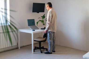 Bir kişi dizüstü bilgisayarı açık bir masanın yanında duruyor. Masanın üzerinde bir bitki var. Odada pencereden gelen parlak bir ışık var..